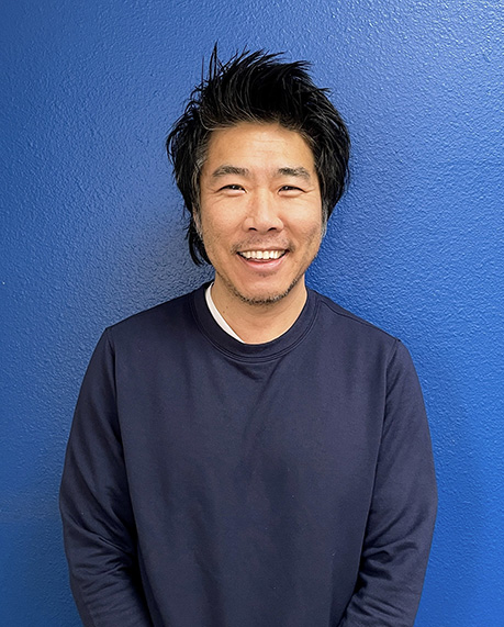 Professional headshot of Director Phil Han smiling at the camera against a bright blue background.
